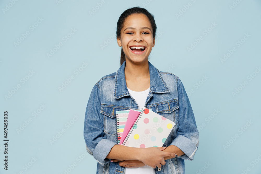 Little smiling fun schoolgirl kid teen girl of African American ethnicity 12-13 years old in denim jacket hold exercisebooks notebooks isolated on pastel plain light blue background Childhood concept Little smiling fun schoolgirl kid teen girl of African American ethnicity 12-13 years old in denim jacket hold exercisebooks notebooks isolated on pastel plain light blue background Childhood concept
