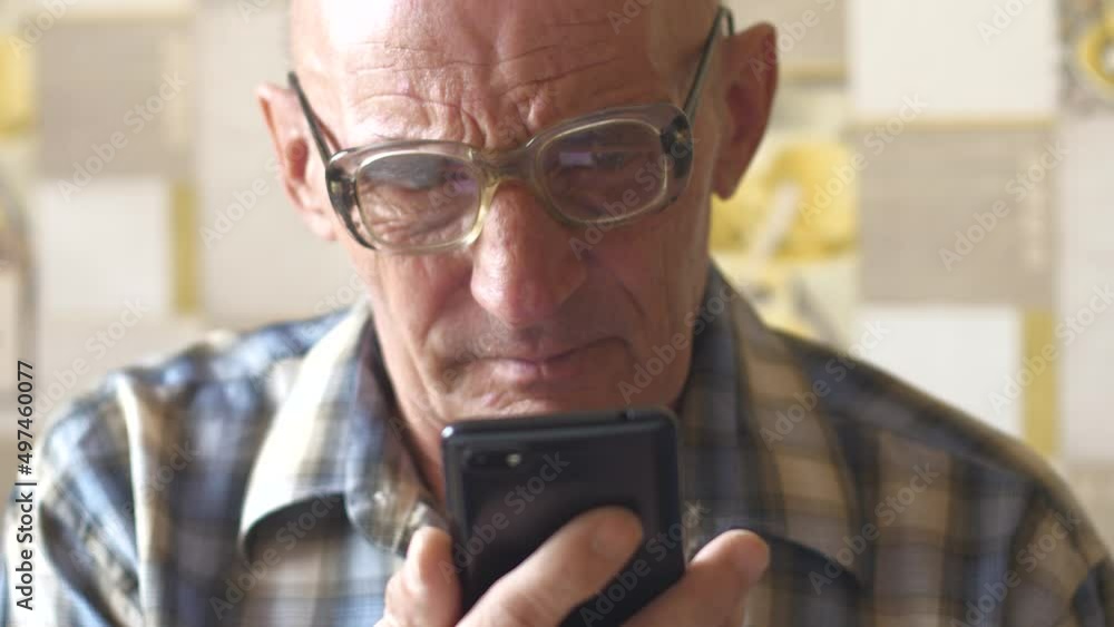 Serious retired caucasian in glasses holds a smartphone and reads information from the screen. Upset elderly man reads a message on the phone. Disturbing news for an old man. selective focus Serious retired caucasian in glasses holds a smartphone and reads information from the screen. Upset elderly man reads a message on the phone. Disturbing news for an old man. selective focus