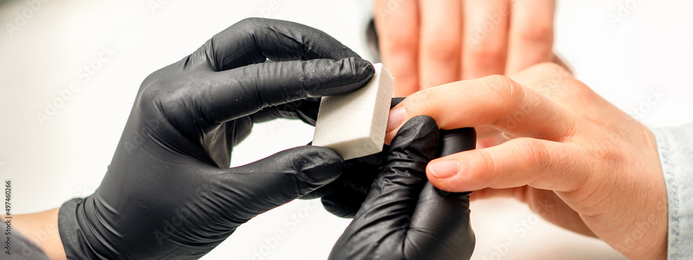 Fototapeta premium Close up of the caucasian hands of a professional manicurist are filing the nails of a young woman. Young caucasian woman receiving a manicure by a beautician with a nail file in a nail salon