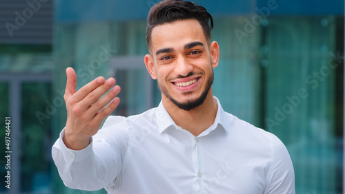 Portrait friendly smiling toothy hispanic arab business man spaniard boss wears white formal shirt showing gesture of invitation advice to approach with hand asks to come outdoors, body languages sign