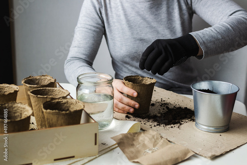 A woman in a gray pullover and one black glove is sitting at a table in the room, scattering plant seeds into peat pots with soil in order to get an environmentally friendly crop after a while.