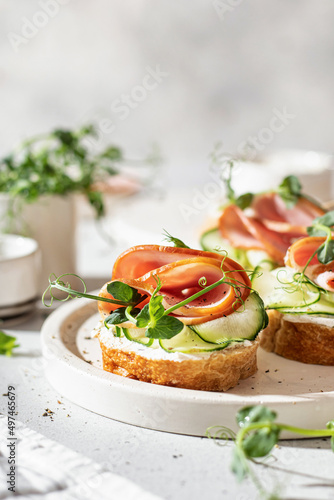 Bruschettas with baguette, bacon or meat, cream cheese, micro-greenery, fresh cucumber and sprouts, in composition on white plate on white textured background