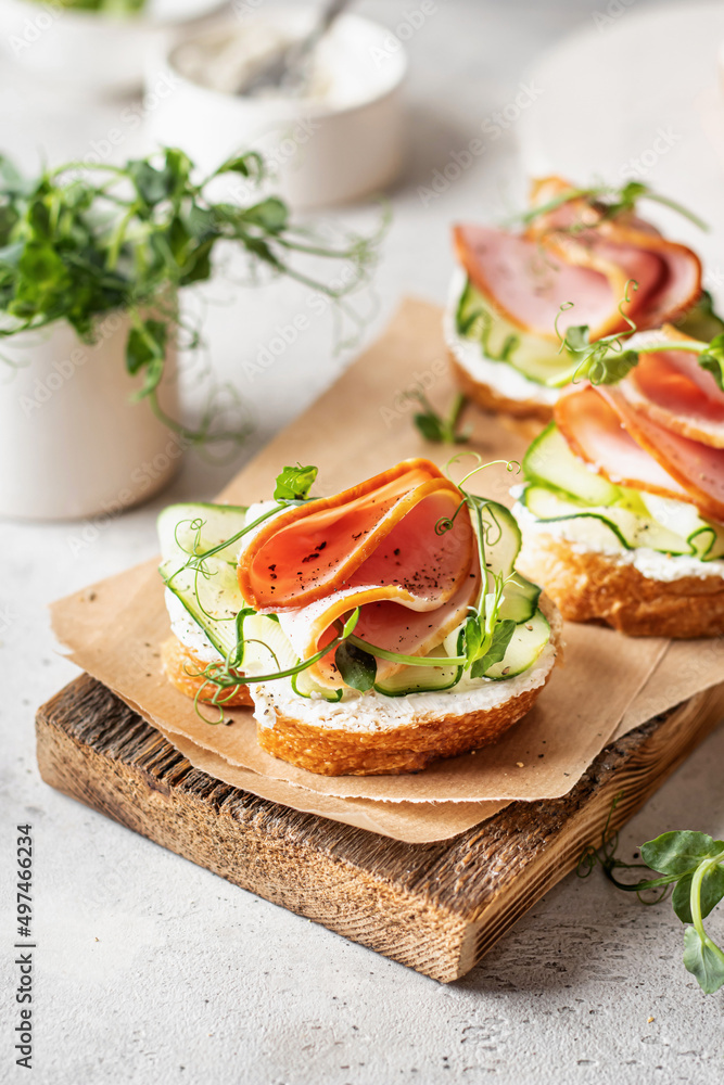 Bruschettas with baguette, bacon or meat, cream cheese, micro-greenery, fresh cucumber and sprouts, in composition on wooden board on white textured background