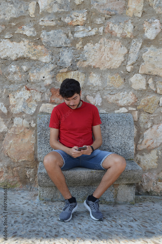Young Caucasian man in red T-shirt looking at his smartphone sitting on a stone bench looking down and copyspace. Young Caucasian man in red T-shirt looking at his smartphone sitting on a stone bench looking down and copyspace.