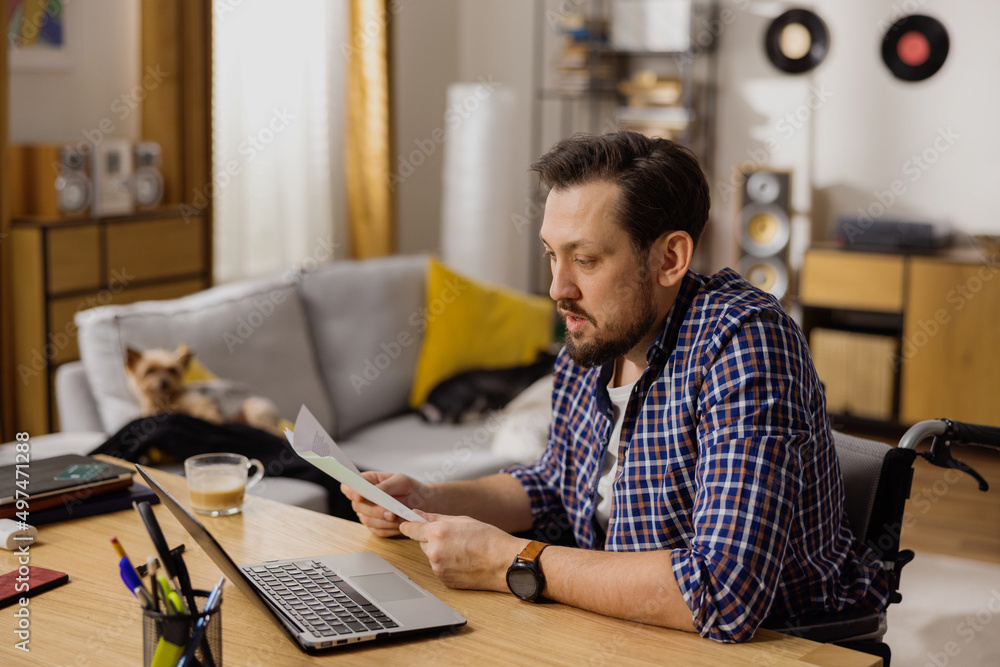 A hard-working man in a wheelchair is at home working on company plans. In his hand he holds documents necessary for his work and in front of him is an open laptop for video conferencing. A hard-working man in a wheelchair is at home working on company plans. In his hand he holds documents necessary for his work and in front of him is an open laptop for video conferencing.