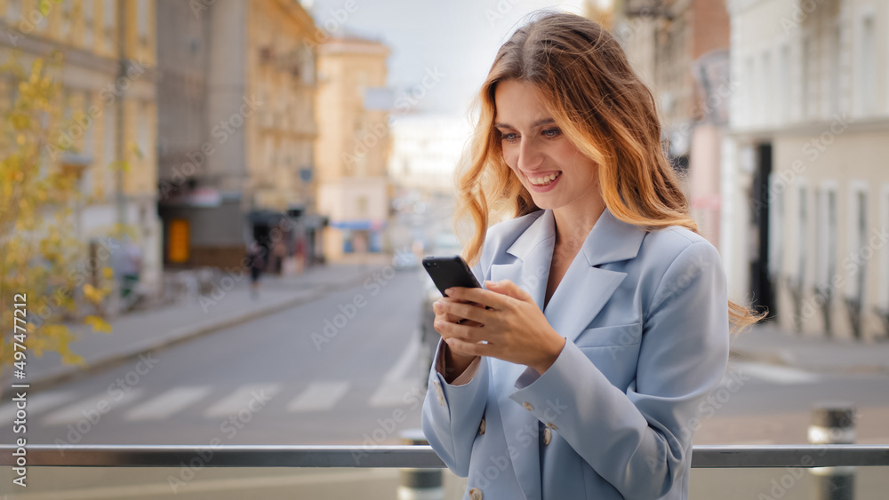 Smiling blonde young girl business woman looking at mobile phone winning rejoices victory triumph makes yes hand gesture shopping online happy with discounts reads good news on balcony city background