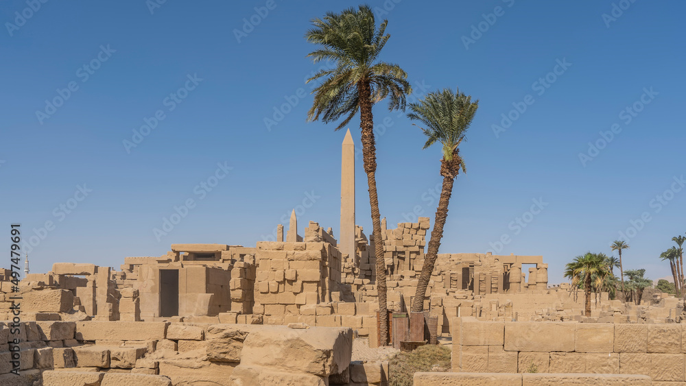 Ruins of the Karnak Temple in Luxor. Dilapidated stone walls and obelisks are visible. Tall palm trees against the blue sky. Egypt