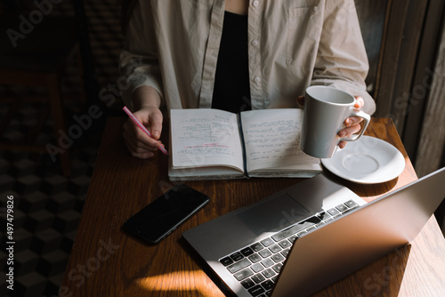 A freelancer girl works in a cafe, drinks cold coffee and makes notes in a notebook with a pink pen; the girl plans her working time