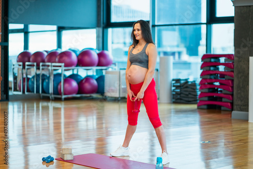 Young dark-haired young pregnant woman dressed in red and grey sportswear and white running shoes squats with dumbbell in gym. Working out and fitness, health care and weight control pregnancy concept