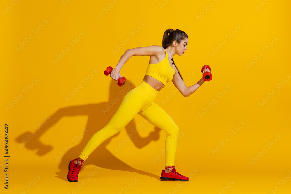 Studio shot of sportive slim girl workout with sports equipment isolated on bright yellow studio background with shadow. Beauty, sport, action, fitness, youth concept.