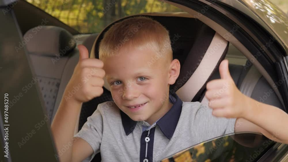 A cute cheerful child is sitting in the car in the back seat, wearing a seat belt and giving a thumbs up. The concept of safe car travel. A cute cheerful child is sitting in the car in the back seat, wearing a seat belt and giving a thumbs up. The concept of safe car travel.