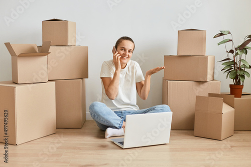 Positive smiling woman wearing t shirt and jeans sitting on floor surrounded with cardboard boxes and talking on phone, having no answer, being not sure, talking with client of real estate agency.