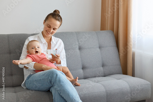 Indoor shot of beautiful Caucasian woman with bun hairstyle wearing white shirt and jeans sitting on sofa with baby and playing, mommy spending time with her toddler child.