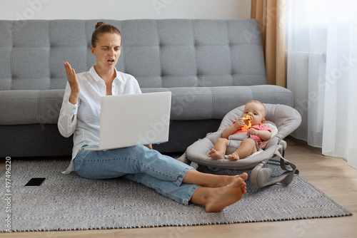 Canvas Print Full length portrait of confused shocked woman wearing white shirt and jeans sitting on floor near sofa with baby in rocking chair, female freelancer looking at screen with puzzlement
