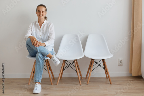 Horizontal shot of smiling positive Caucasian woman wearing white shirt and jeans sitting on chair in queue, expressing confidence, looking at camera with smile.