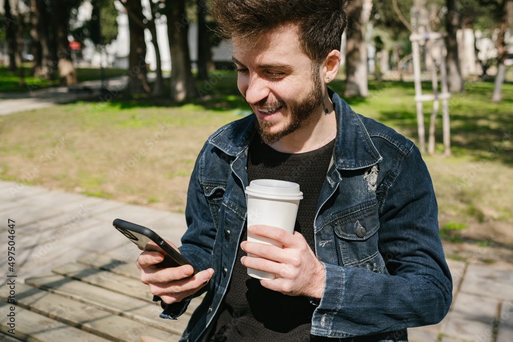 Young happy man looking phone screen while holding a cup of coffee at outside. Man breathing fresh air at the outdoor while drinking coffee. He feel relax and happy with healthy lifestyle. Young happy man looking phone screen while holding a cup of coffee at outside. Man breathing fresh air at the outdoor while drinking coffee. He feel relax and happy with healthy lifestyle.