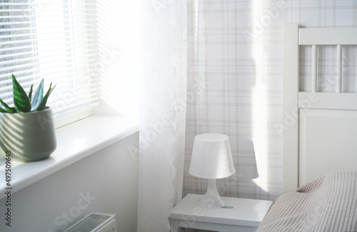 The interior of the room in white tones with a bed, bedside table, table lamp and a flower on the windowsill © Viktoriia