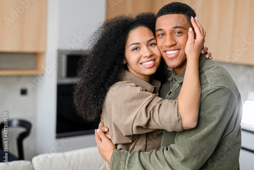 Lovely multiracial couple in embraces in cozy modern kitchen at home. Smiling guy is hugging charming girl. Love and affection concept