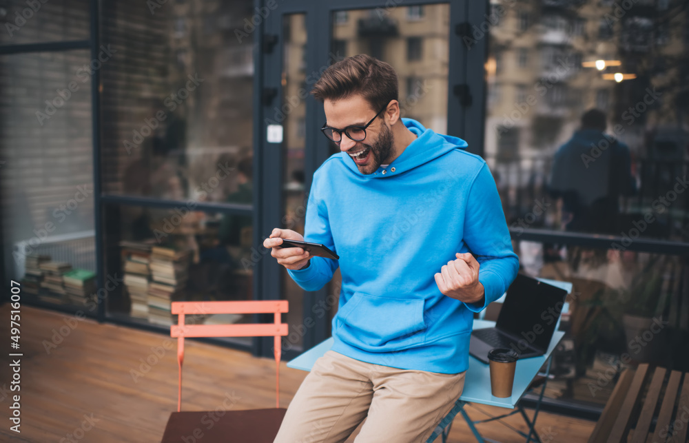 Joyful hipster guy in classic spectacles for provide vision correction celebrating win in online sports betting using cellphone gadget for checking received results, excited Caucasian male blogger Joyful hipster guy in classic spectacles for provide vision correction celebrating win in online sports betting using cellphone gadget for checking received results, excited Caucasian male blogger