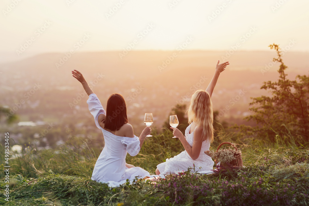 Two young happy female with hands up in white summer dresses at picnic. Carefree women making picnic at sunset.