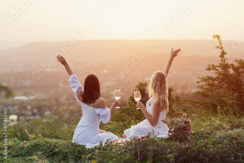 Obraz na płótnie Two young happy female with hands up in white summer dresses at picnic. Carefree women making picnic at sunset. w salonie