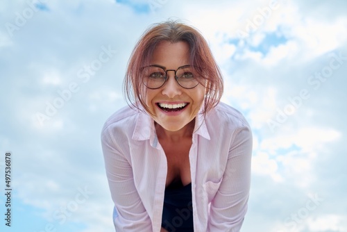 Happy middle age woman looking down at camera, blue sky in clouds background.