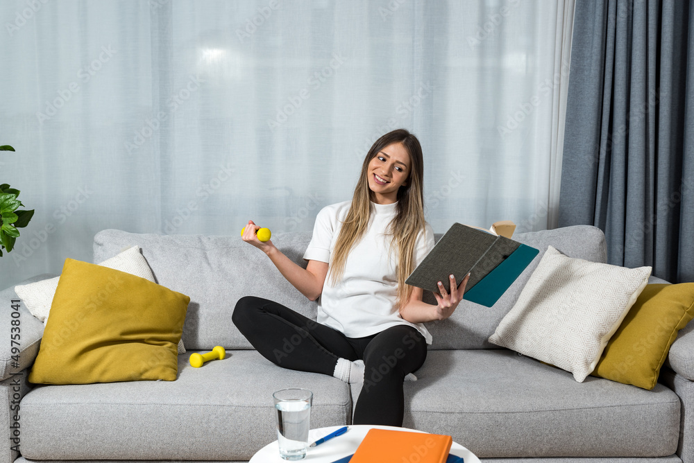 Young happy fitness college student woman sitting at home study reading books to educate herself holding small weight and exercise with one hand. Health and motivated life concept Young happy fitness college student woman sitting at home study reading books to educate herself holding small weight and exercise with one hand. Health and motivated life concept