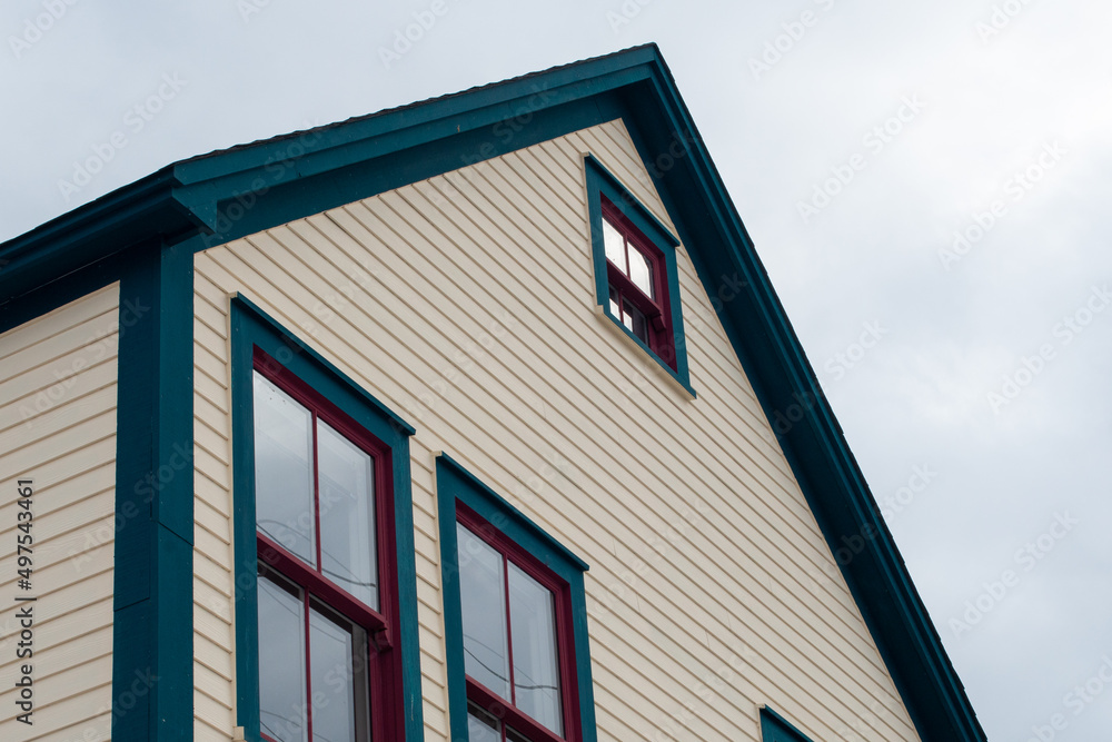 The upward view of the exterior roof section of a wooden vintage building. The colorful yellow, green and red painted house has multiple single hung windows reflecting the cloudy blue sky. 