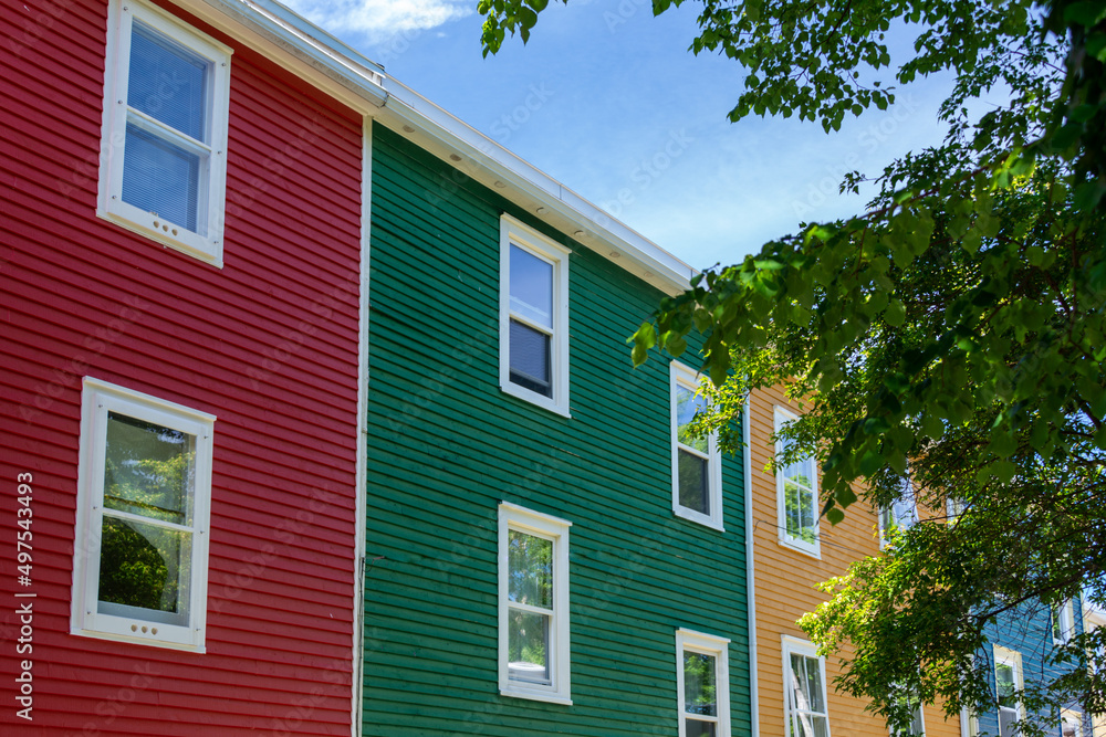 An upward view of an exterior of multiple joined colorful wooden houses with double hung windows. There's a red, green, yellow, and blue adjacent building. There's a blue sky and a large green tree. 
