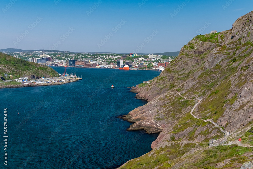 A footpath, hiking trail, or path along a hillside. The cliff is rocky with grass patches. The city of St. John's, Newfoundland, is in the background on a sunny day. The sky is bright blue. 