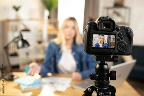 Smart blond young business lady in casual clothes filming educational lecture on video camera while sitting at table. Focus on camera screen. Blur background.