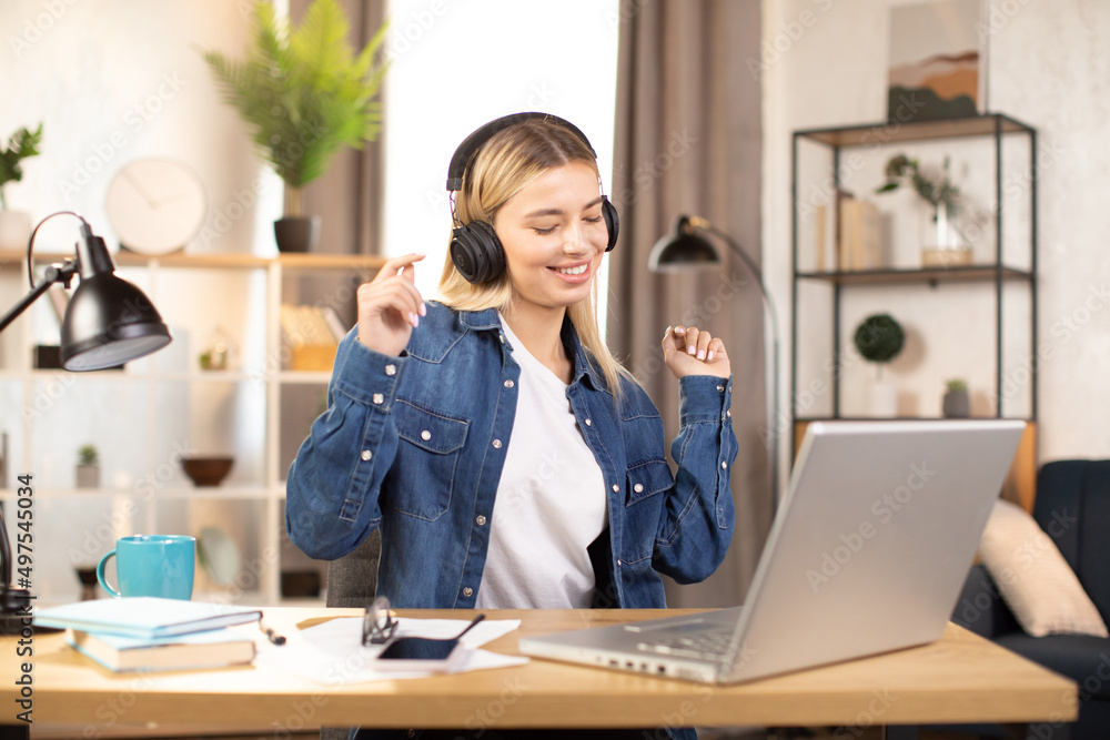 Portrait of a beautiful young blonde woman dressed in casual jeans shirt, listening musing with headphones and dancing. Relaxation during work. Break after study. Remote, freelance