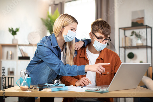 Students, man and woman in casual outfit and face masks looking at computer screen during working meeting. Young partners cooperating or working remotely online from home