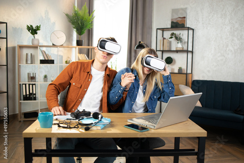 Excited young people with surprised expression and open mouth, playing games at home, using VR glasses and laptops. Happy family enjoying artificial reality entertaining themselves with innovations.