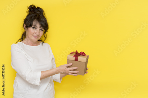 beautiful aged woman holds out a gift box on a yellow background with a place for text, the concept of giving a gift