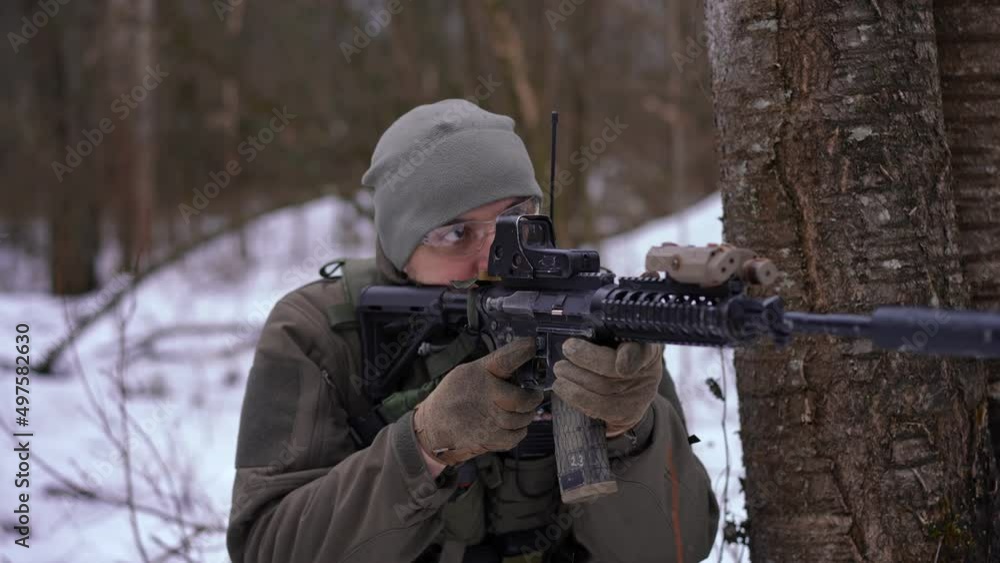 Portrait of courageous young man with gun walking in winter forest sitting down at tree aiming with weapon. Brave Ukrainian male soldier outdoors in war zone. Military intelligence concept Portrait of courageous young man with gun walking in winter forest sitting down at tree aiming with weapon. Brave Ukrainian male soldier outdoors in war zone. Military intelligence concept