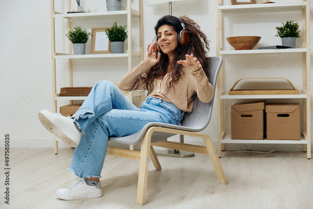 Relaxed calm pleasant tanned curly Latin lady with headphones moves to cool music sit on chair in home modern interior close eyes. Copy space Mockup Banner. Full body shoot. Relax in home Relaxed calm pleasant tanned curly Latin lady with headphones moves to cool music sit on chair in home modern interior close eyes. Copy space Mockup Banner. Full body shoot. Relax in home
