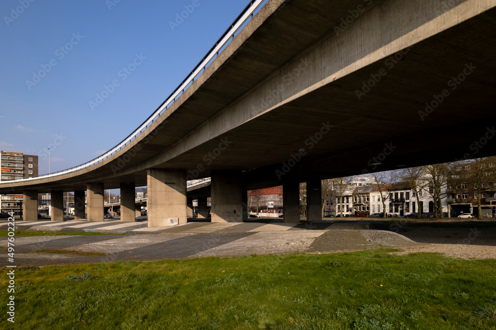 Fototapeta premium Overpass Arnhem intersection Onderlangs roundabout seen from below the concrete construction with apartment buildings in the background. Transportation and infrastructure engineering concept.