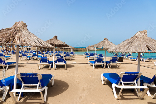 Empty beach with chairs, sunbeds, straw umbrellas, and blue sky. Mediterranean summer vacation destination, Heraklion, Crete, Greece.