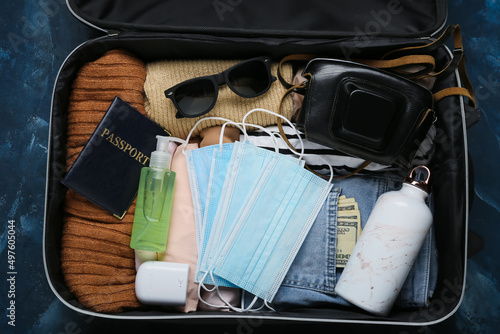 Suitcase with stylish female clothes, accessories, medical masks and earphones on dark background