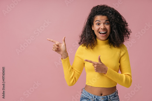 Excited African American woman wearing yellow sweater pointing finger on copy space isolated on pink background. Advertisement concept © ARUTA Images