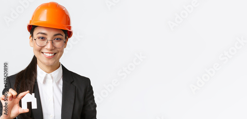 Close-up of happy asian female engingeer, real estate agent in helmet and business suit holding house miniature and smiling, architect working over renovation project, white background photo