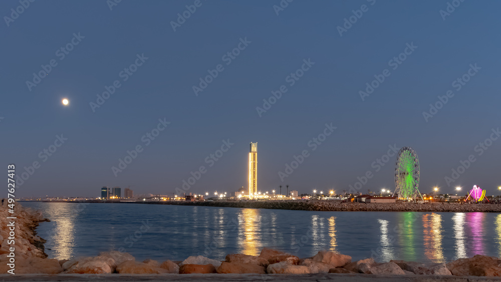 Fototapeta premium The great mosque and ferris wheel under moonlight seen from Sablette promenade jetty. Algiers city by night, water reflexion of city lights illuminated by colorful world biggest minaret.