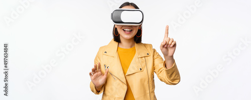 Image of young asian woman wearng VR headset, using virtual reality glasses and smiling, pointing at smth, choosing smth, standing over white background photo