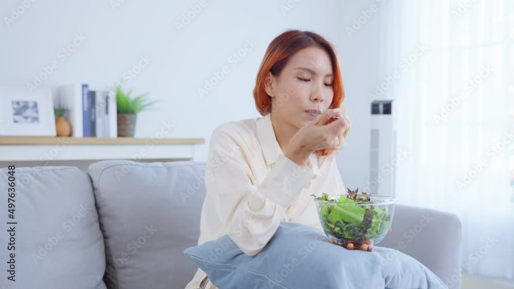 Asian attractive woman sit on sofa, eating green salad in living room. Asian attractive woman sit on sofa, eating green salad in living room.