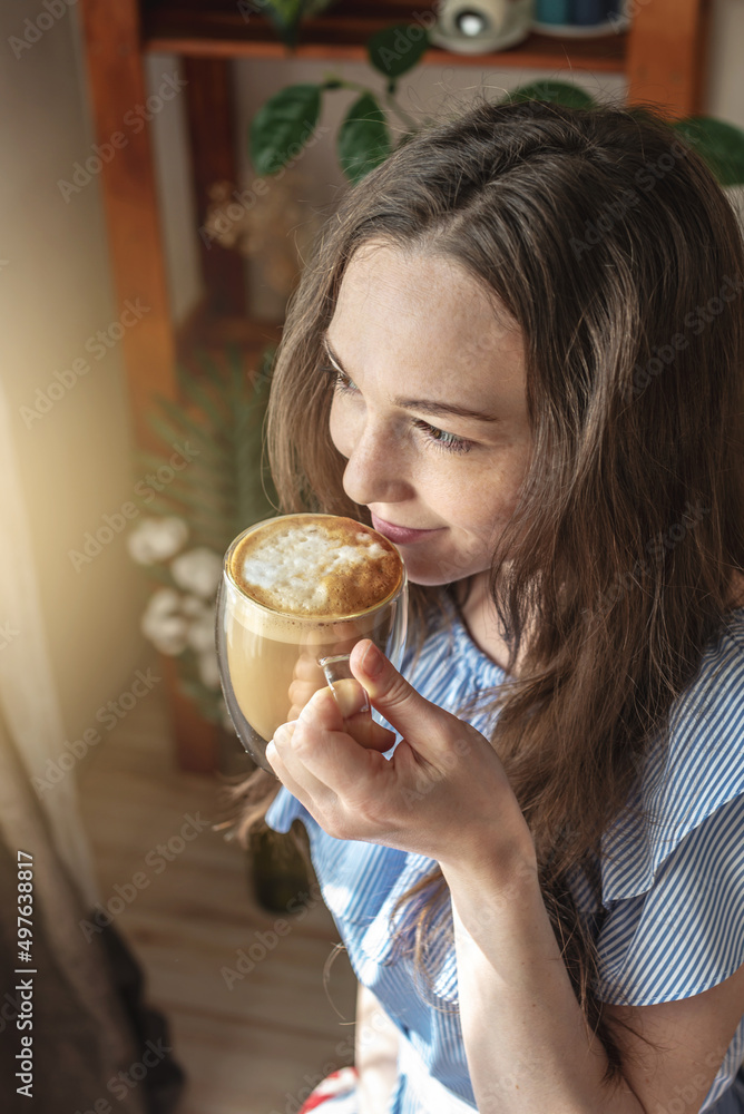 Young pretty woman is sitting near the window and enjoying a fragrant morning coffee. Concept of a pleasant pastime, good mood and happiness in small stuff Young pretty woman is sitting near the window and enjoying a fragrant morning coffee. Concept of a pleasant pastime, good mood and happiness in small stuff