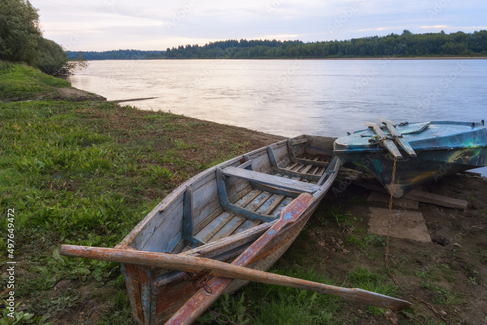 On the bank of the river, old boats with oars are waiting for their fishermen for early summer fishing (Ural, Russia). The sky and water are illuminated by the rising sun, the forest on the banks
