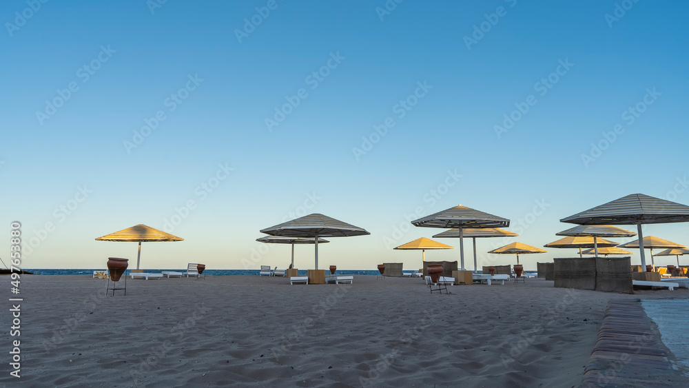 Morning on the Red Sea coast. The sun illuminates the umbrellas. Sunbeds and ceramic urns stand in the shade on the sand. Clear blue sky. Egypt. Safaga