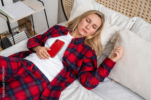 A young woman is lying on a bed in pajamas, sleeping with a mobile phone on her chest. The concept of a good mood before going to bed.