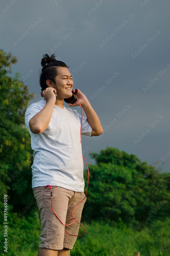 asian male model handsome smile wearing t-shirt and casual short listening to music song using headphone while squinting his eyes from the glare of the light asian male model handsome smile wearing t-shirt and casual short listening to music song using headphone while squinting his eyes from the glare of the light